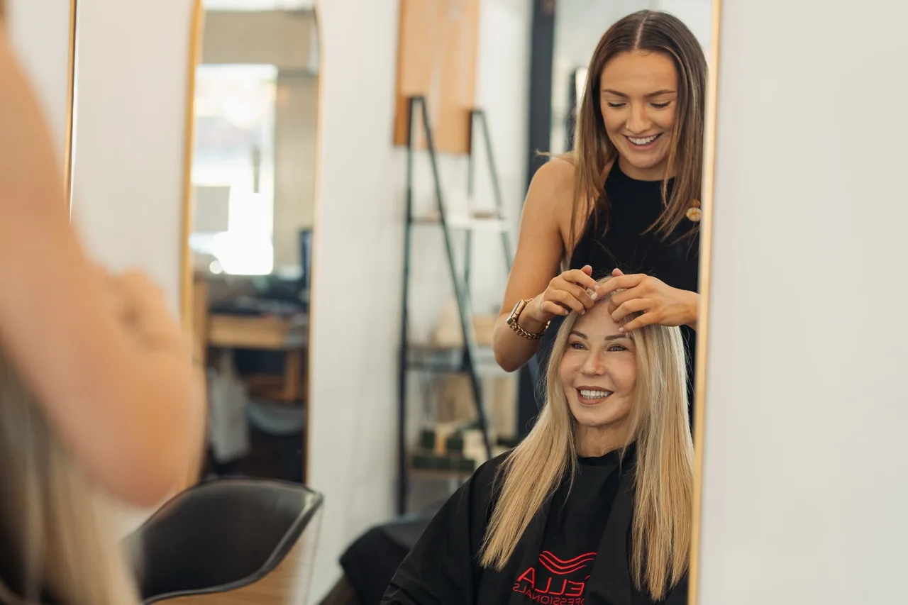A smiling stylist standing behind a seated blonde-haired client at a salon mirror
